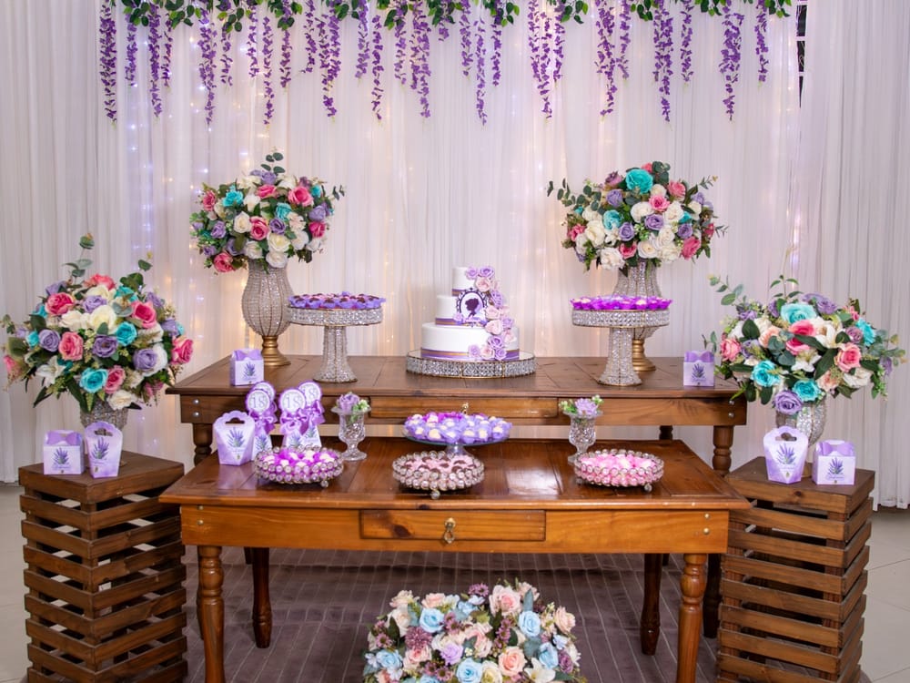 Mesa de bolo com arranjos de rosas lilás, rosa e azul, glicínias lilás e cortina de luzes